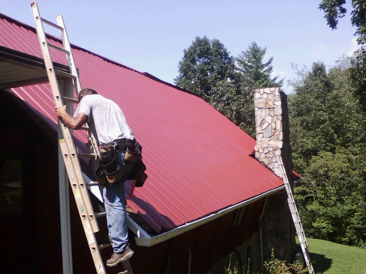 Roofer on a ladder beside a red metal roof and stone chimney — Soffit & Fascia Repair by Roofing Brothers in Bethel Park